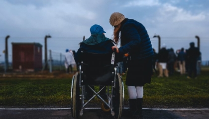Due persone di spalle, una ragazza in piedi che parla con l'altra seduta su una sedia a ruote e guardano in avanti, in un luogo all'aperto