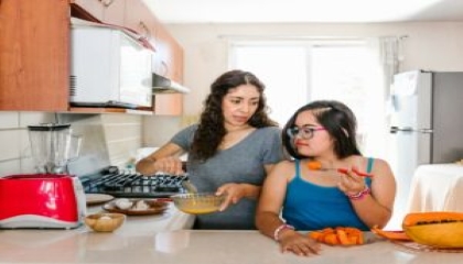 All’interno di una cucina, una donna adulta spiega come cucinare una pietanza ad una ragazza con sindrome di Down, mentre quest’ultima mangia del melone poggiata sul piano di lavoro.